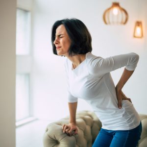 Woman makes painful face while leaning on furniture and grasping her lower back