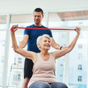 Woman sitting on a yoga ball does an overhead stretch with a resistance band while being assisted by a smiling man