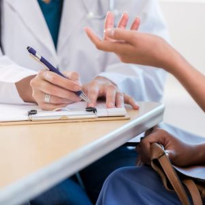 We see two people sitting at a table, but just their hands. One person is a doctor, going over some papers on a clipboard. The other is a woman holding a purse on her lap, gesturing with her hands.