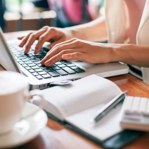 WOman's hands typing on a laptop. Her desk is cluttered with a coffee cup, notebook, and calculator