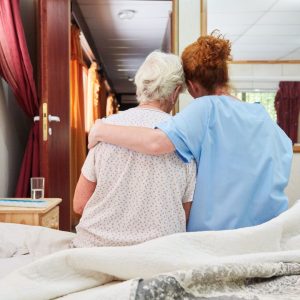 Two women sit on a hospital bed with their backs towards us. A younger woman in scrubs has put her arm around an older woman, who is leaning into the embrace in comfort.