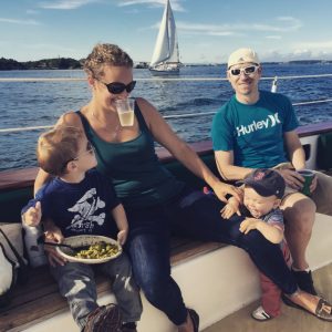 Family on a sailboat; the mom has white wine in a plastic cup held by her teeth while she juggles two toddlers and the dad smiles