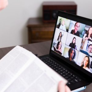 A laptop is open with people's faces on the screen, like a zoom call. Someone has a book open in front of the lap top
