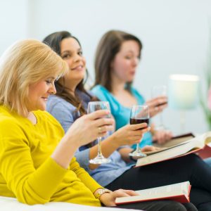 A group of women sit in chairs with books open on their laps, and glasses of red wine in hand.