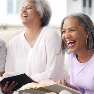 Two women are sitting together on a couch and laughing while holding books open in their laps