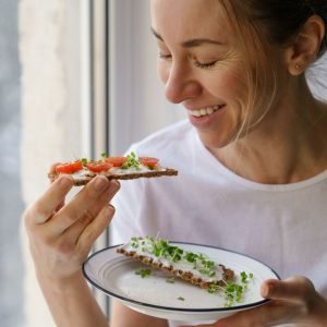 Fresh-faced woman wearing a white t-shirt smiles as she lifts a plate with food towards her. She is holding a flatbread snack with fresh veggies on top