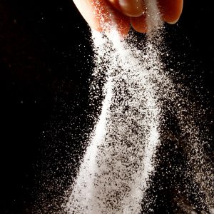 Dramatically lit fingers sprinkle a cascade of salt in front of a black background.