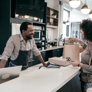 Man wearing an apron and standing behind a counter smiles at a customer, who is paying for something in a brown paper bag