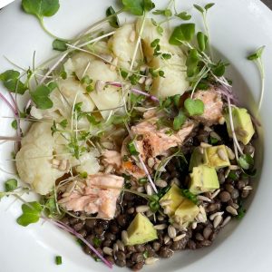 Aesthetically pleasing arrangement of lentils, cut up avocado, chunks of salmon, cauliflower, and sprouts in a white bowl set on a dark wooden table