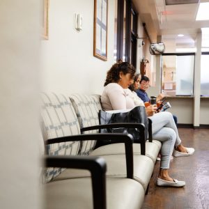 We can see several people sitting in chairs against the wall of a waiting room, looking at their phones