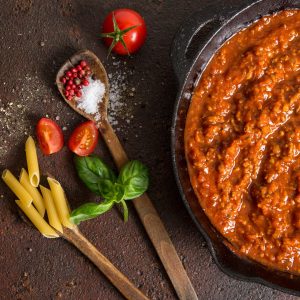 A pot of meat sauce simmers in a cast-iron skillet, while next to it rests a wooden spoon, scattered salt, tomatoes, and pasta
