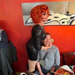 A drag queen with bright red hair holds a man's head to her bosom. The man is seated and smiling with his eyes closed