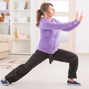 A woman practices tai chi at home in her living room, wearing a purple sweater and gray, loose-fitting pants