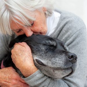 A grey-haired woman hugs the head of a grey-muzzled black dog. Both of them have closed their eyes; the dog looks serene, the woman looks sad.