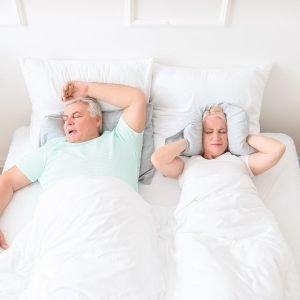 In an overwhelmingly white bedroom, an older couple is asleep in bed. The man is snoring with his arm over his head, and the wife is clutching a pillow over her ears.