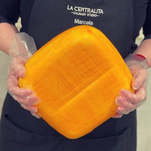 A woman wearing a black apron with the logo for La Centralita Culinary Studio is holding a huge rounded-square block of cheese