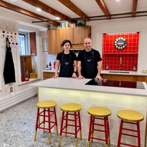 A young couple dressed in black and wearing black aprons stand proudly behind a cooktop counter.