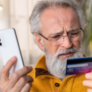 A man with grey hair and beard frowns at his credit card while holding a cell phone to his ear