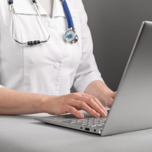 A female doctor wearing a white lab coat and a stethoscope around her neck is typing on a laptop