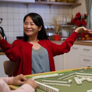 Woman triumphantly raises her hands and looks smugly at the people she is playing mahjong with at the kitchen table