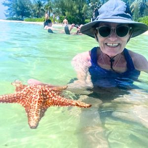 A woman wearing a floppy beach hat smiles as she holds up a starfish while sitting in the water