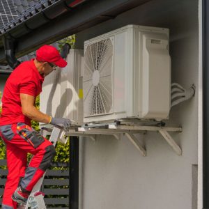 A man wearing all red inspects an airconditioning unit on the side of a building while perched on a ladder