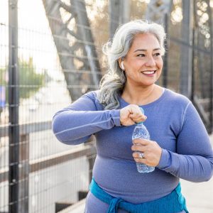 A woman with long, grey hair smiles as she uncaps a bottle of water during a walk