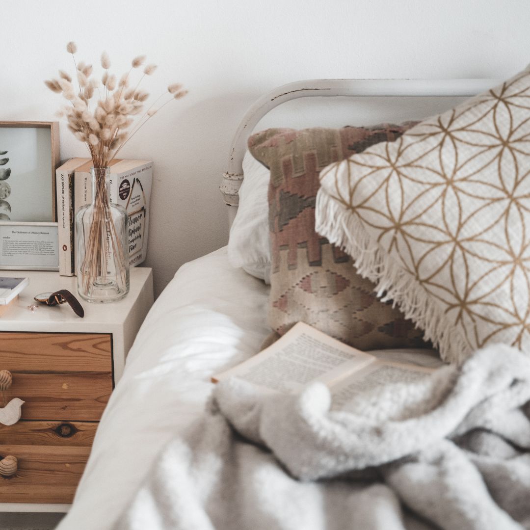 a cozy, slept-in looking bed has a soft, grey fleece blanket and several textured throw pillows in neutral hues. On the bedside table there is a small vase with dried grasses, and books