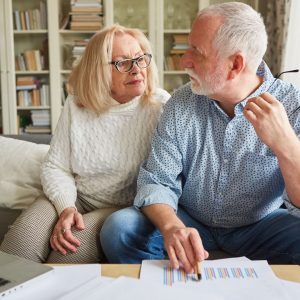 An older couple sit on the couch and go over financial papers. They're looking at each other seriously.