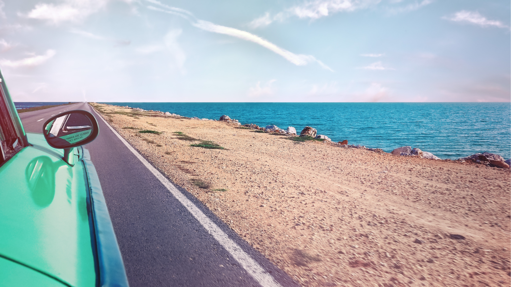 View from a passenger side seat of a teal green car, driving down a road that follows a narrow, sandy coastline